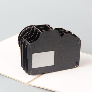May include: A black and gray 3D pop-up card shaped like a camera. The card is open and the camera is standing up. The camera has a black and gray textured surface and a silver lens.