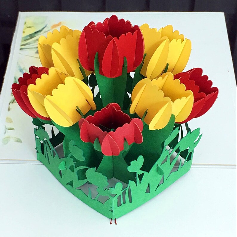 May include: A 3D pop-up card featuring a bouquet of red and yellow tulips with green leaves and stems. The card is made of paper and is designed to stand upright.