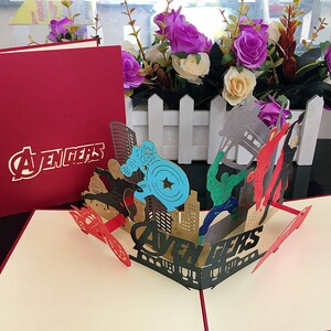 May include: A red and cream pop-up card featuring the Avengers characters. The card is open and shows a 3D scene of the Avengers characters in a cityscape. The card has the word "Avengers" printed on the front and inside.
