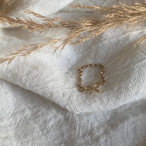 May include: A gold chain ring with a delicate bow charm. The ring is on a white fabric background.