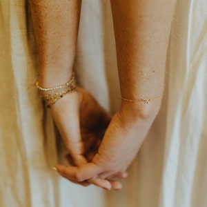 May include: Two wrists with delicate gold bracelets. One bracelet has a small, sparkling charm. The bracelets are on a person's arms, but the person's hands are visible.