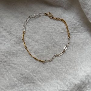 May include: A delicate bracelet featuring a mix of gold and silver chain links. The bracelet has a clasp closure and is displayed on a white, textured surface. The design incorporates various link styles, creating a two-tone effect.