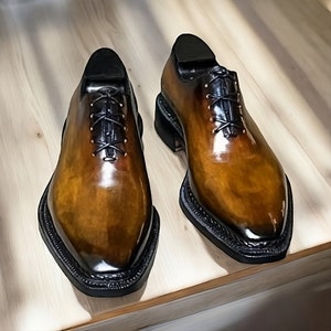 Puede incluir: Un par de zapatos de cuero marr&oacute;n con cordones, suelas y cordones negros. Los zapatos tienen una punta puntiaguda y un acabado pulido.