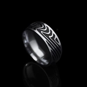 Könnte beinhalten: Ein silberner Ring mit einem schwarzen, wellenartigen Muster um das Band. Der Ring ist hochglanzpoliert und reflektiert den dunklen Hintergrund. Das Design ist symmetrisch und modern. Die Ringbreite beträgt ca. 0,8 cm.