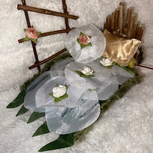 May include: A miniature bed with a white and green floral bedspread, a gold pillow, and a white pillow with a pink rose. The bed is made of twigs and is surrounded by green moss.