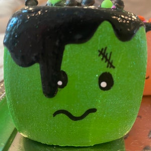 May include: Green and black Halloween-themed cake pop with a Frankenstein face and black icing dripping down the side. The cake pop is decorated with white eyes and a stitched scar.