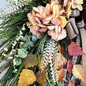 May include: A decorative wreath featuring peach-colored hydrangea blossoms, green fern fronds, white berry sprigs, and autumn leaves in shades of yellow, orange, and red. The arrangement includes brown textured ribbon accents and a woven twig base.