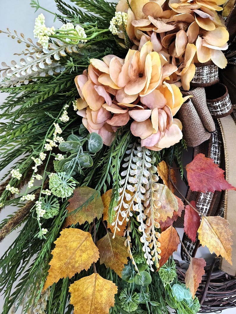 May include: A decorative wreath featuring peach-colored hydrangea blossoms, green fern fronds, white berry sprigs, and autumn leaves in shades of yellow, orange, and red. The arrangement includes brown textured ribbon accents and a woven twig base.