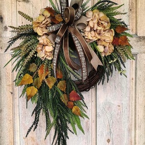 May include: A decorative wreath featuring a brown twig base adorned with beige hydrangea blossoms, green fern-like foliage, and scattered autumn-colored leaves in shades of yellow, orange, and brown, finished with a brown and plaid ribbon bow.