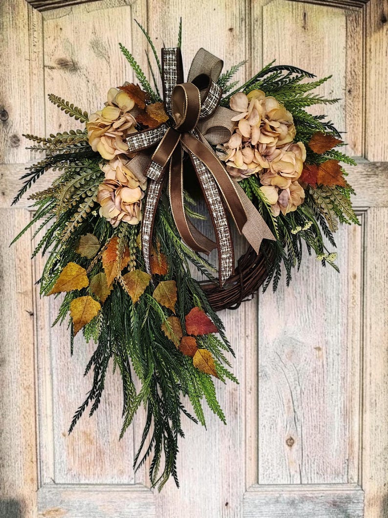 May include: A decorative wreath featuring a brown twig base adorned with beige hydrangea blossoms, green fern-like foliage, and scattered autumn-colored leaves in shades of yellow, orange, and brown, finished with a brown and plaid ribbon bow.