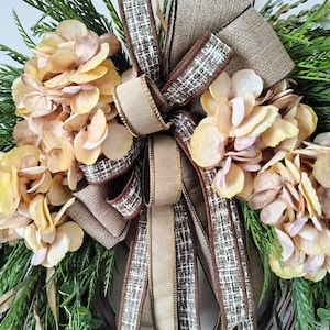 May include: A decorative wreath featuring clusters of light peach hydrangea flowers and green foliage arranged on a brown twig base. A large, multi-layered bow made of tan burlap and brown-and-white checkered fabric is centered on the wreath.