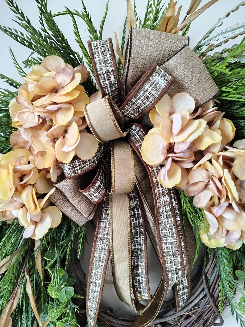 May include: A decorative wreath featuring clusters of light peach hydrangea flowers and green foliage arranged on a brown twig base. A large, multi-layered bow made of tan burlap and brown-and-white checkered fabric is centered on the wreath.