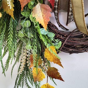 May include: A decorative wreath featuring a dark brown woven twig base adorned with artificial autumn foliage, including orange, yellow, and brown serrated leaves, green fern-like sprigs, small round green berries, and a brown and gold ribbon.