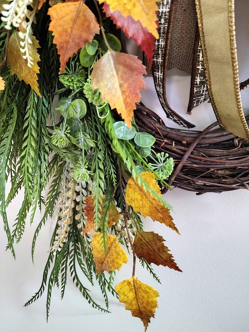 May include: A decorative wreath featuring a dark brown woven twig base adorned with artificial autumn foliage, including orange, yellow, and brown serrated leaves, green fern-like sprigs, small round green berries, and a brown and gold ribbon.