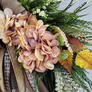 May include: A decorative wreath featuring muted peach and pink hydrangea blossoms, green foliage, and yellow autumn leaves. The arrangement is accented with long, trailing brown and tan textured ribbons and small white floral sprigs on a twig base.