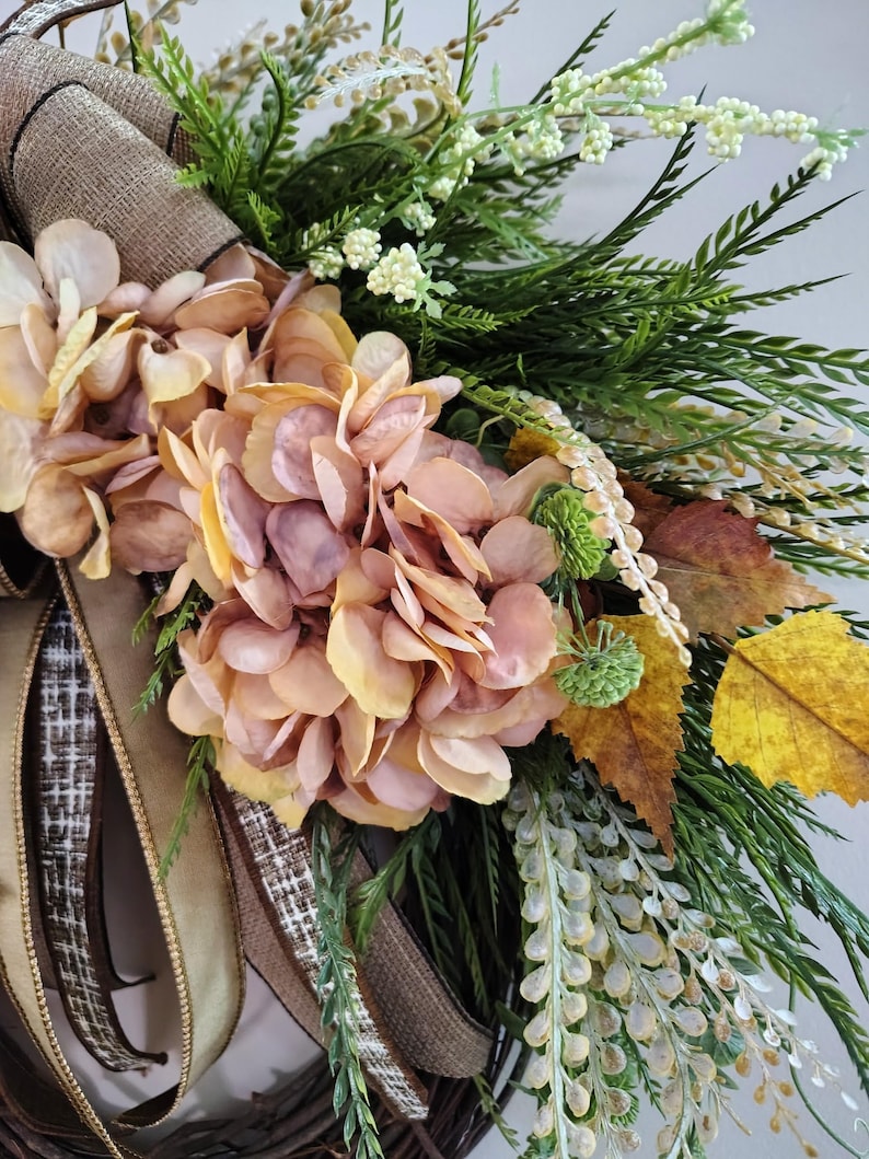 May include: A decorative wreath featuring muted peach and pink hydrangea blossoms, green foliage, and yellow autumn leaves. The arrangement is accented with long, trailing brown and tan textured ribbons and small white floral sprigs on a twig base.