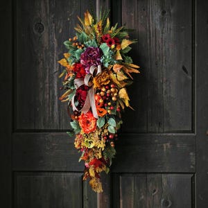 May include: A teardrop-shaped autumn floral swag featuring a mix of artificial orange, red, and deep purple flowers, yellow and brown foliage, and clusters of small berries. A light beige ribbon is tied in a bow at the center of the arrangement.