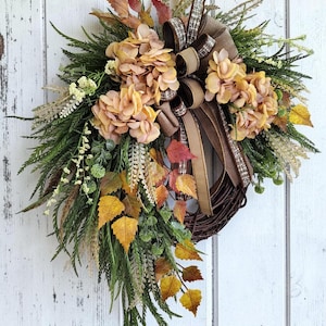 May include: A teardrop-shaped wreath featuring a dark brown twig base adorned with light orange hydrangea blooms, green fern fronds, yellow and orange autumn leaves, and a decorative bow made of brown and tan plaid and solid fabric ribbons.