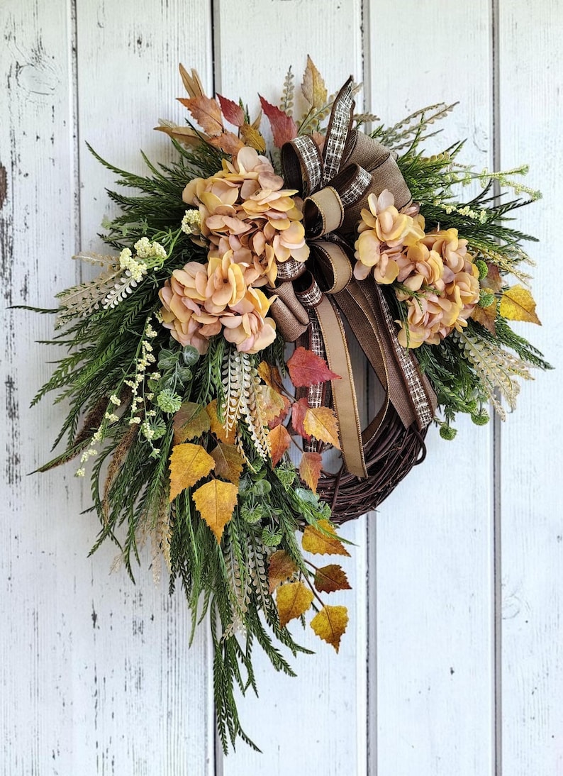 May include: A teardrop-shaped wreath featuring a dark brown twig base adorned with light orange hydrangea blooms, green fern fronds, yellow and orange autumn leaves, and a decorative bow made of brown and tan plaid and solid fabric ribbons.