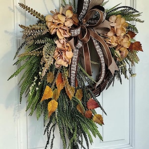 May include: A teardrop-shaped wreath featuring a dark brown twig base adorned with green fern fronds, dried beige hydrangea blossoms, and autumn-colored leaves in shades of yellow, orange, and red, finished with a brown and white plaid ribbon bow.