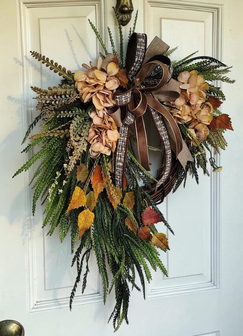 May include: A teardrop-shaped wreath featuring a dark brown twig base adorned with green fern fronds, dried beige hydrangea blossoms, and autumn-colored leaves in shades of yellow, orange, and red, finished with a brown and white plaid ribbon bow.