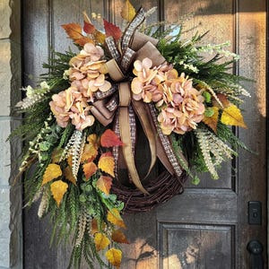 May include: A decorative wreath featuring a dark brown twig base adorned with artificial green pine needles, yellow and orange autumn leaves, and clusters of light peach hydrangea flowers, finished with a brown and plaid fabric ribbon bow.