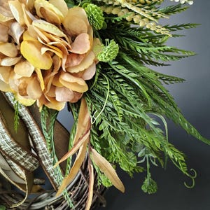 May include: A decorative wreath featuring large, golden-yellow hydrangea blossoms, vibrant green fern fronds, and small green flower buds. The arrangement is secured to a natural brown twig base and accented with a textured, tan-colored ribbon.