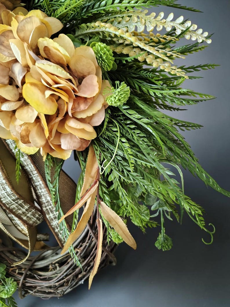 May include: A decorative wreath featuring large, golden-yellow hydrangea blossoms, vibrant green fern fronds, and small green flower buds. The arrangement is secured to a natural brown twig base and accented with a textured, tan-colored ribbon.