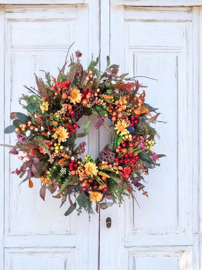 May include: A fall wreath with orange, red, and yellow flowers and berries. The wreath is made of artificial greenery and is hanging on a white door.