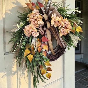 May include: A decorative wreath featuring a dark brown twig base adorned with peach-colored hydrangea blossoms, green pine-like foliage, and autumn-toned leaves in yellow and orange. A multi-layered brown and plaid ribbon bow is centered at the top.