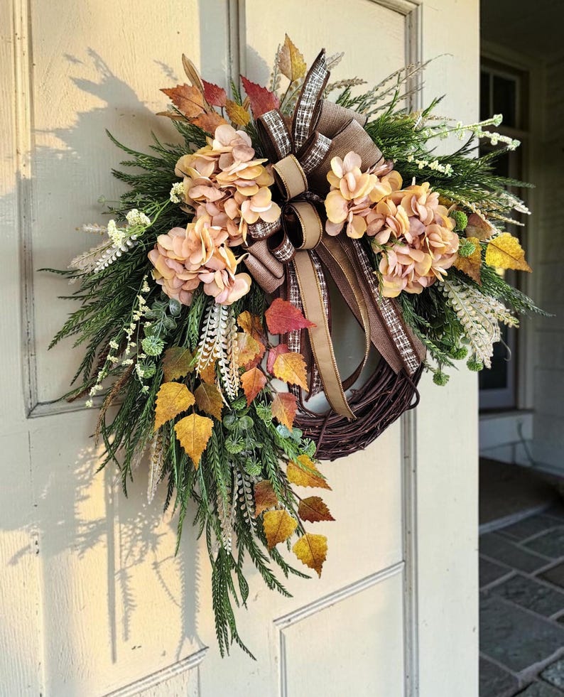 May include: A decorative wreath featuring a dark brown twig base adorned with peach-colored hydrangea blossoms, green pine-like foliage, and autumn-toned leaves in yellow and orange. A multi-layered brown and plaid ribbon bow is centered at the top.
