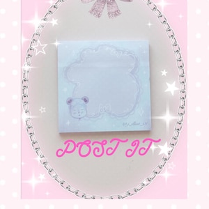 May include: A square Post-it note with a light blue cloud-shaped design and a cute bear illustration. The text "POST IT" is written in pink cursive. The image is framed with a silver chain and pink bow.