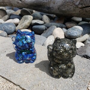 Puede incluir: Dos figuras de gato de resina. Una es azul con inclusiones coloridas, la otra es negra con motas doradas. Ambas están en posición sentada con una pata levantada, sobre una superficie de hormigón, con un fondo natural.