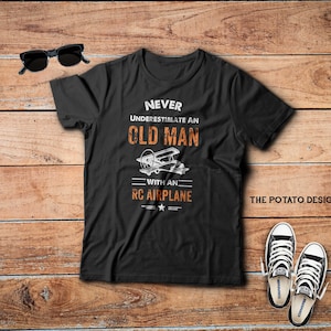 May include: Black t-shirt with white text that reads "Never underestimate an old man with an RC airplane". The text is in a distressed font. There is a small airplane graphic above the text. The words "The Potato Design" are in white text on the bottom right corner of the shirt.