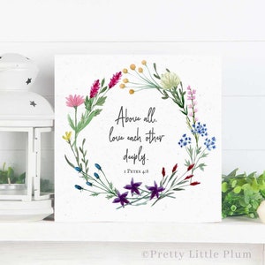 Puede incluir: Una impresión en lienzo cuadrado blanco con una corona de flores y el texto "Above all, love each other deeply. 1 Peter 4:8". La corona está hecha de flores de acuarela en tonos de rosa, morado, azul y amarillo.
