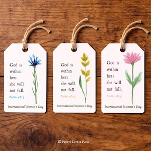 May include: Three white gift tags with watercolor floral designs and the text "God is within her; she will not fall. Psalm 46:5. International Women's Day." Each tag has a twine string.