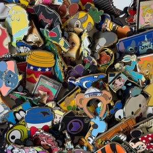 Peut inclure: Une grande collection d'épinglettes en émail métallique coloré représentant divers personnages de dessins animés, y compris des figures Disney, des tasses à café et des icônes thématiques, disposées en un tas dense et superposé.