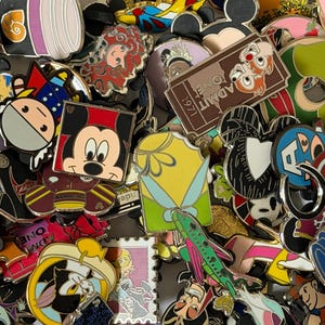 Op de afbeelding: Een grote verzameling diverse metalen emaille pins met verschillende stripfiguren, filmiconen en symbolen. De pins bevatten ontwerpen van Mickey Mouse, Tinker Bell, een bruin ticket met de tekst ADMIT ONE 1971 en een groen vliegtuig.