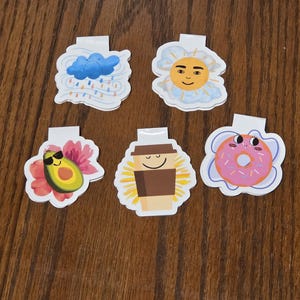 May include: Set of six colorful, illustrated paper clips. Designs include a cloud with rain, a smiling sun, an avocado, a coffee cup, and a pink donut. Each clip has a white tab.