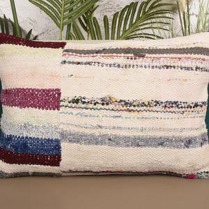 May include: Rectangular decorative pillow with a patchwork design. The pillow features a cream-colored base with sections of pink, purple, blue, and burgundy. The pillow is made of woven fabric and has a textured appearance.