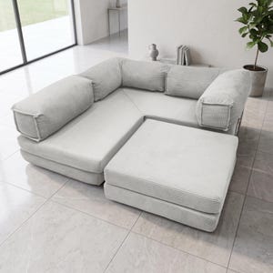 May include: A light gray modular sofa set featuring a textured corduroy fabric. The configuration includes a corner section with backrests and a separate square ottoman piece, all designed with clean lines and a low-profile, boxy silhouette.
