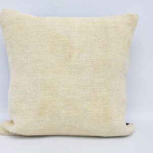 May include: A square throw pillow featuring a textured, off-white, woven fabric cover. The material displays a visible, coarse, and uniform weave pattern throughout the entire surface, creating a neutral and solid appearance for this home decor item.