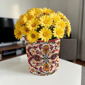 Op de afbeelding: Een boeket gele madeliefjes in een decoratieve vaas. De vaas heeft een gedetailleerd, kleurrijk patroon met rode, blauwe en gouden accenten. De bovenrand van de vaas is rood afgewerkt.