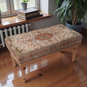 Peut inclure: Un banc rectangulaire avec un dessus à motifs rappelant un tapis ancien. Le motif du tapis présente un médaillon central dans des tons orange et rouge, entouré de motifs floraux et géométriques. Le banc a des pieds en bois clair.