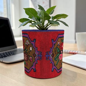 May include: A red plant pot with a detailed, patterned design in brown, blue, and green. The pot holds a green leafy plant. The pot is cylindrical and sits on a wooden surface, with a laptop and a mug in the background.