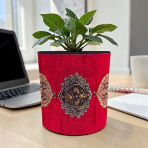 Può includere: Un vaso rosso con un motivo ornamentale nei toni del marrone, nero e oro. Il vaso contiene una pianta verde con foglie. Il vaso cilindrico è appoggiato su una scrivania di legno, con un laptop e una tazza sullo sfondo.