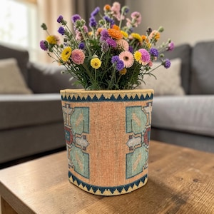 May include: A cylindrical flower pot with a patterned design in shades of peach, teal, and navy blue, filled with a vibrant assortment of colourful flowers. The pot sits on a wooden table, showcasing a blend of textures and hues.