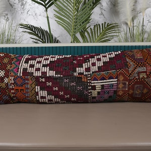 May include: A long, rectangular pillow with a patchwork design. The pillow features a variety of colors, including burgundy, brown, white, and orange. The pillow is approximately 30 inches long and 12 inches wide, with a decorative, textured surface.