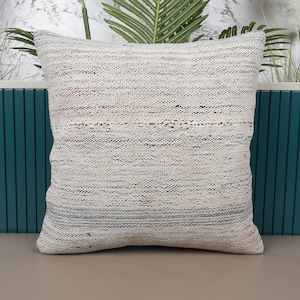 May include: A square, textured pillow in shades of white, gray, and a hint of pink. The pillow has a woven appearance and is placed against a teal-colored backdrop. The pillow is a decorative item for a home.