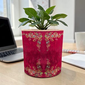 Op de afbeelding: Een cilindervormige, bordeauxrode bloempot met een bloemenontwerp. De pot heeft een gedetailleerd patroon van kleurrijke bloemen en bladeren. De pot bevat een groene plant met bladeren, wat een levendig contrast creëert. De pot is waarschijnlijk van stof.